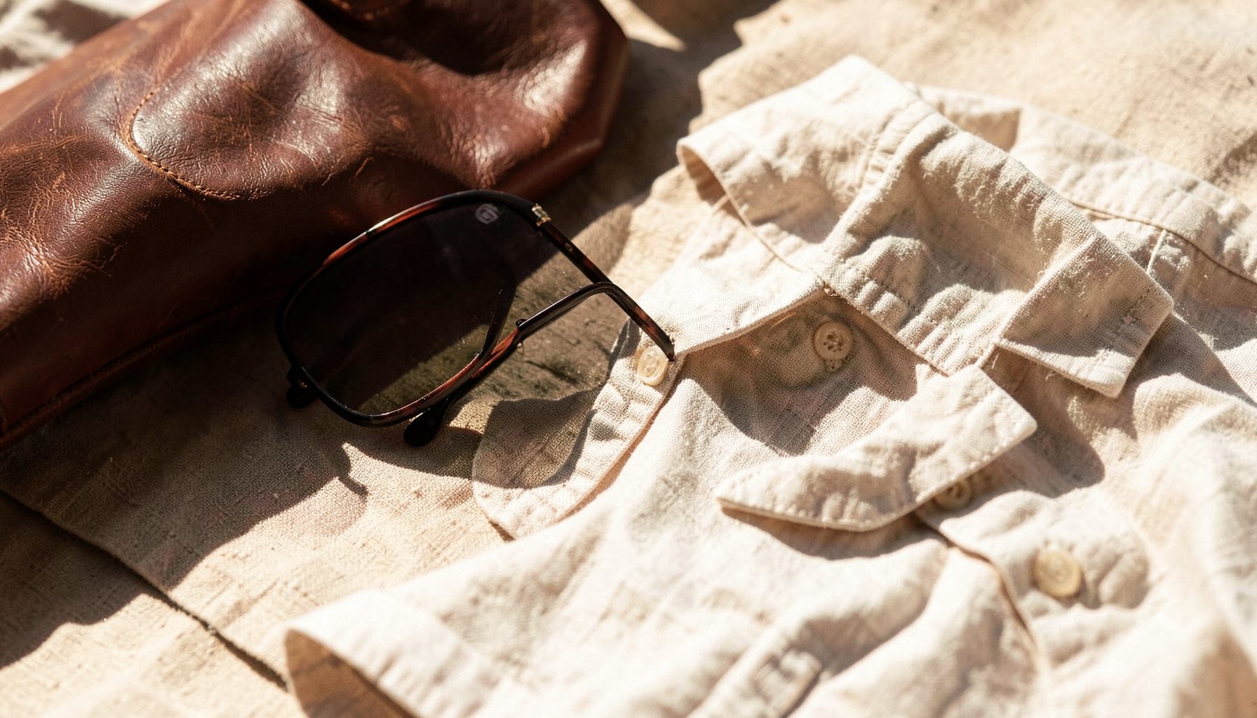 Warm-toned lifestyle scene with sunglasses, a leather bag, and a linen shirt on a sunlit surface